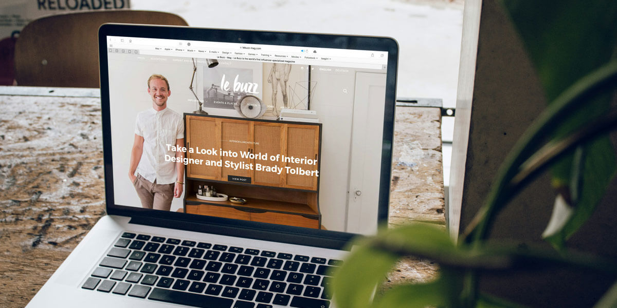 A laptop on a wooden table displays a website featuring an interior designer and stylist, with a plant partially visible in the foreground.